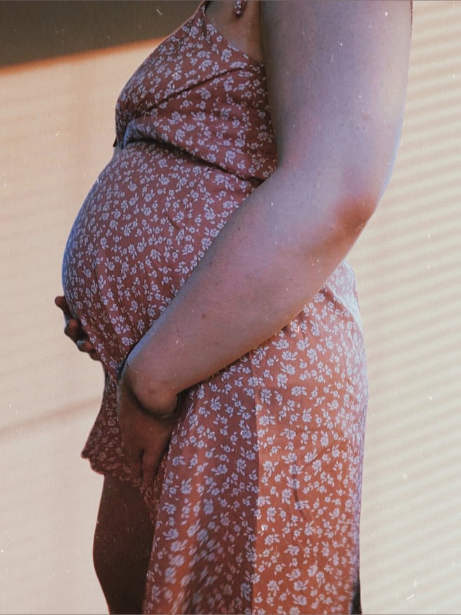A pregnant woman in a floral-patterned dress standing against a beige wall in natural light.