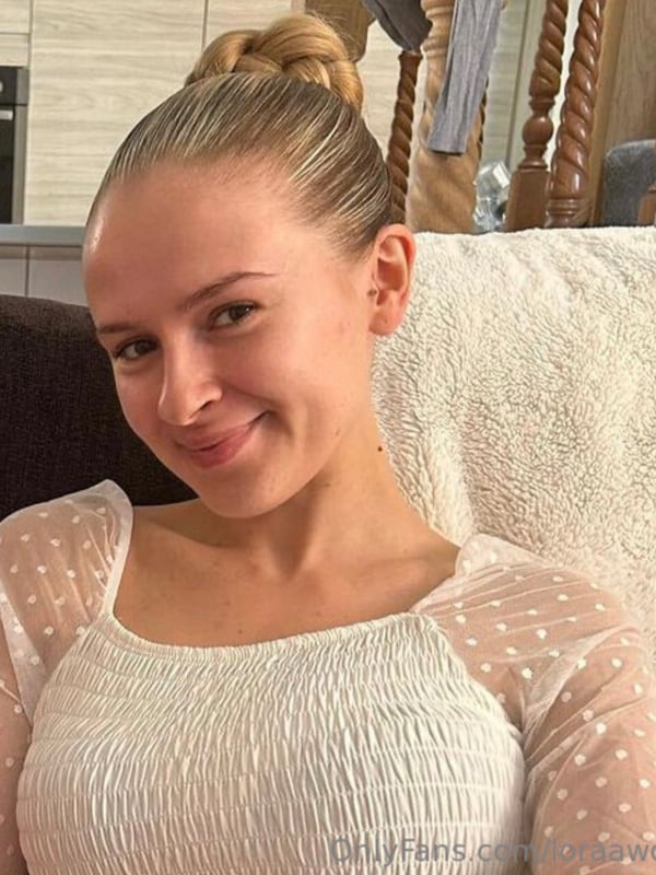 A smiling young woman with blonde hair in a bun, sitting in a kitchen wearing a sheer white top.