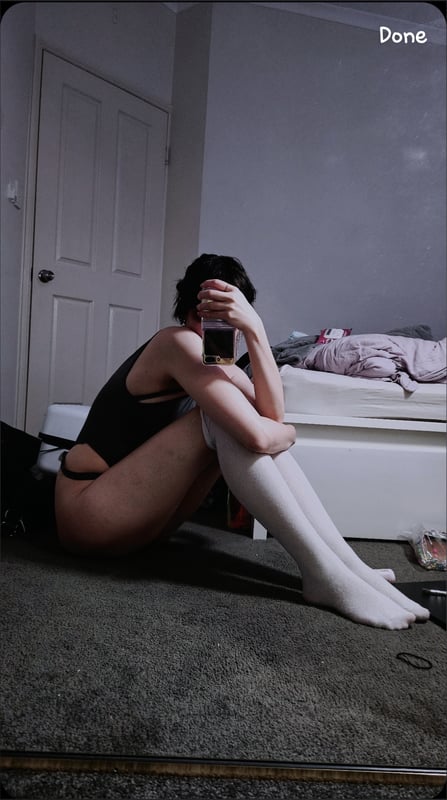 A person sitting on the floor in a dimly lit bedroom, wearing a black tank top and white thigh-high socks, with hands covering their face.