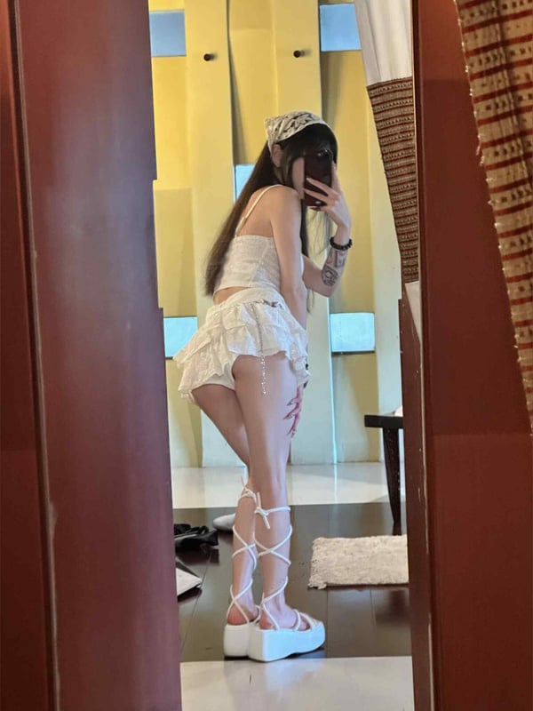 Young woman in white crop top and mini skirt with platform sandals posing in front of a mirror in an indoor setting with warm lighting.