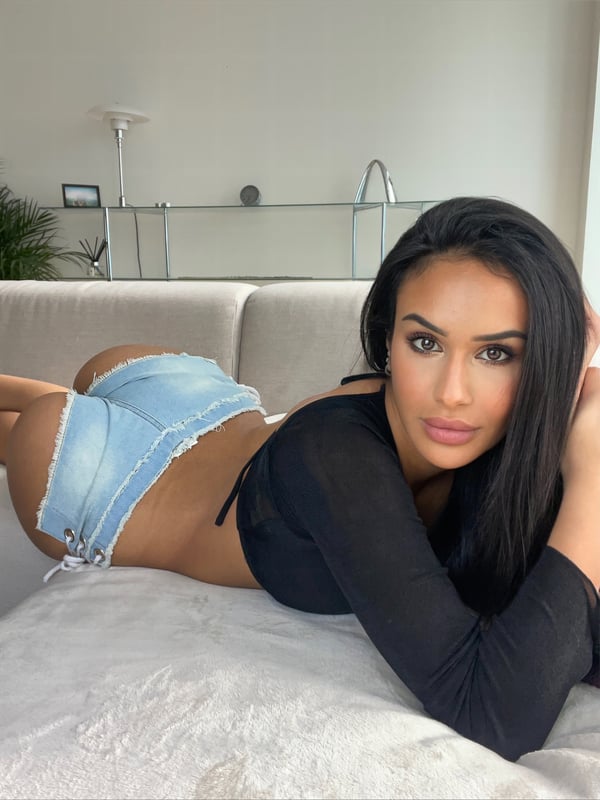 A woman with long dark hair lying on a couch in a modern indoor setting, wearing a black top and light blue shorts.