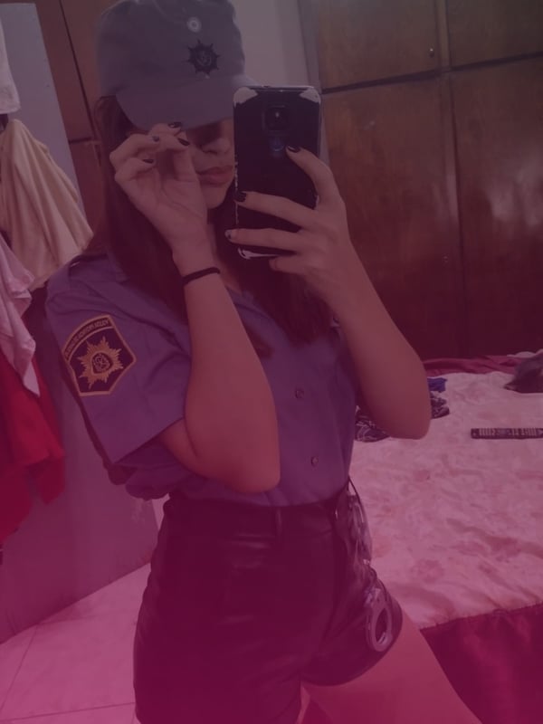 Young woman wearing a police uniform shirt and shorts, tipping a cap while taking a mirror selfie in a bedroom.