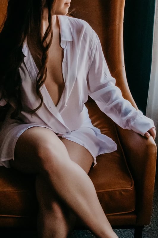 A woman with long dark hair sits on a brown leather armchair, wearing a loose white shirt, with soft lighting near a window.