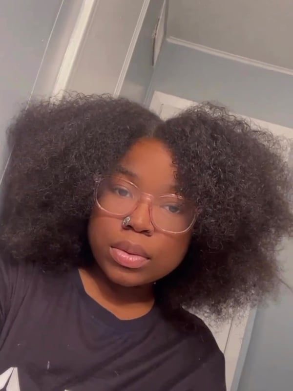 A young woman with curly black hair wearing glasses and a black T-shirt, smiling at the camera indoors.