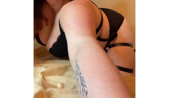 A woman posing on a bed in black lingerie with garter straps, showing tattoos on her arm.