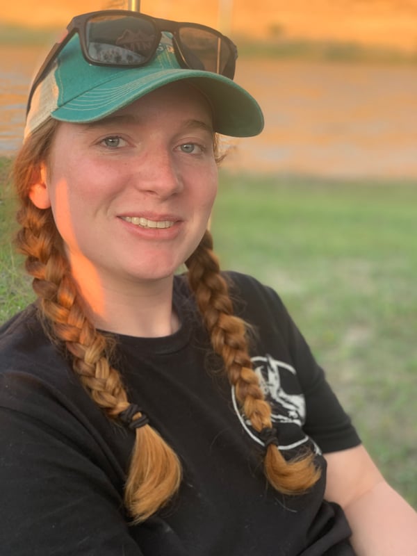 Young woman with red hair in braids, wearing a green cap and sunglasses, smiling outdoors in a black t-shirt against a sunset background.