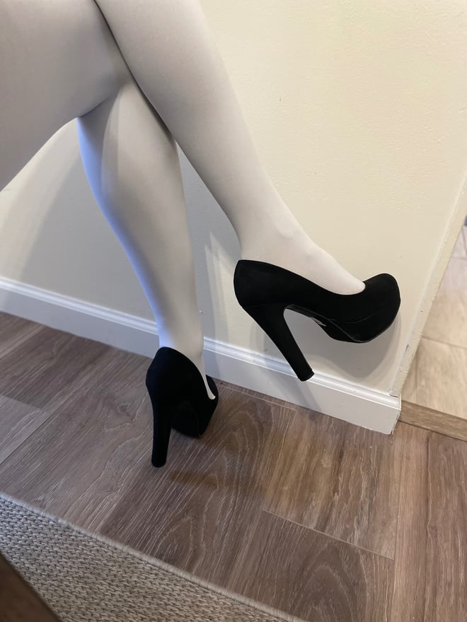 Close-up of a woman's legs in white pantyhose and black high heels positioned on a floor against a plain wall.