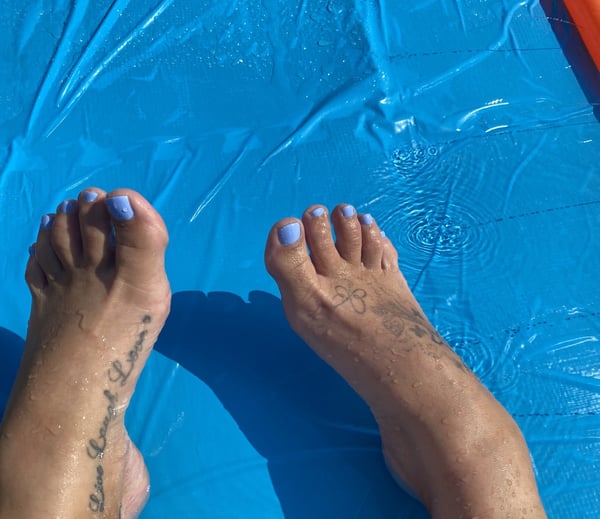 Close-up view of feet with light blue nail polish and tattoos, positioned on a blue inflatable surface with water droplets.