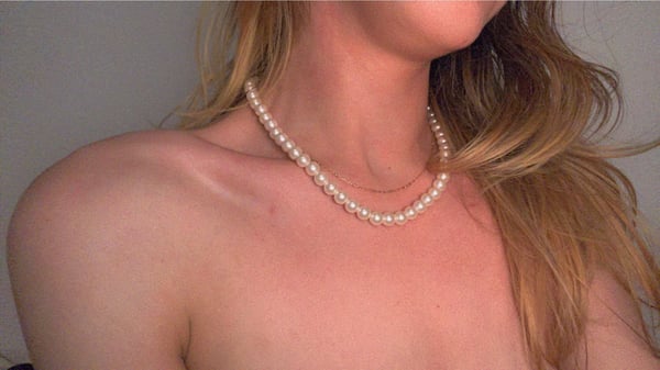 A young woman with long blonde hair wearing a pearl necklace, posing with shoulders visible against a plain background.