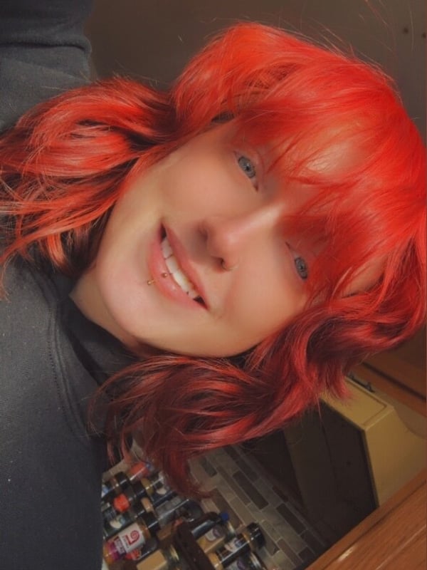 Young woman with bright red hair smiling at the camera, standing in front of a shelf of bottles in a kitchen.