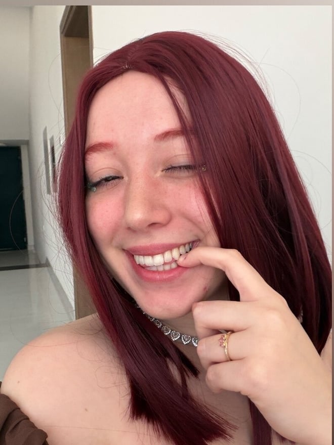 A young woman with long red hair smiling with her hand near her mouth, wearing a choker necklace, against a plain indoor background.