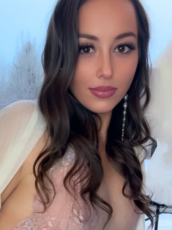 A woman with long dark wavy hair smiling confidently outdoors against a snowy landscape, wearing a sheer pink off-shoulder top.