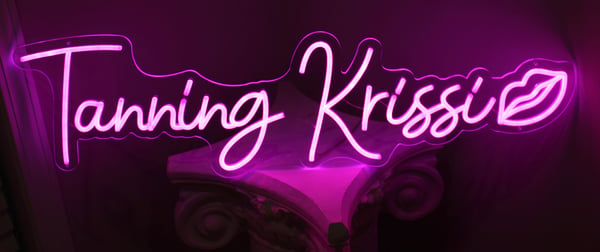 Neon pink sign displaying 'Tanning Krissi' with a kiss mark, illuminated against a dark background.