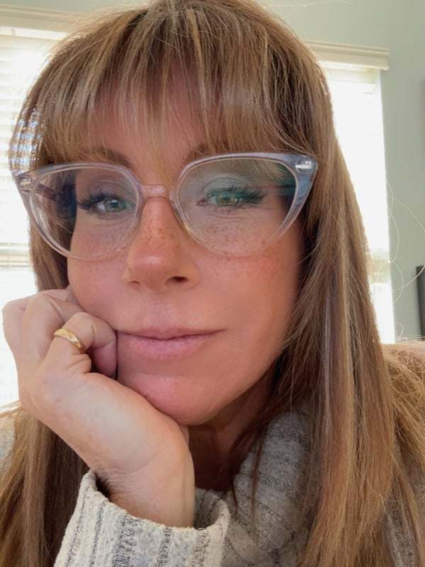 A woman with blonde hair and glasses smiling softly while resting her chin on her hand, dressed in a cozy sweater indoors.