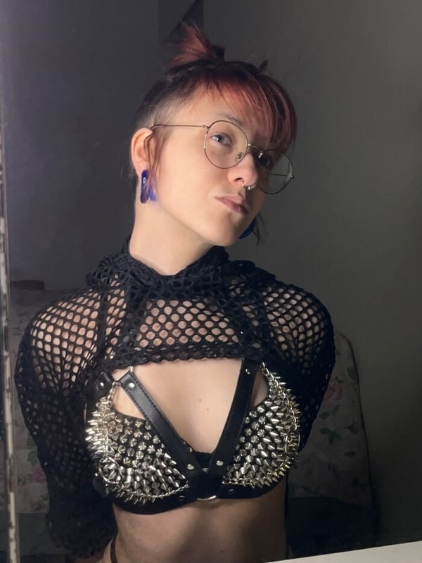 A person with short pinkish hair and glasses wearing a studded black mesh top and hoodie, posing confidently indoors.