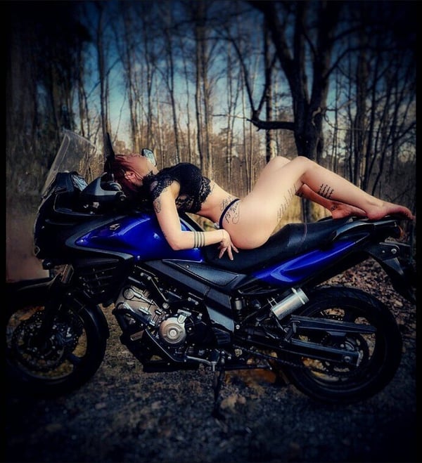 A woman in black lingerie reclining on a blue motorcycle in a forested area during twilight.