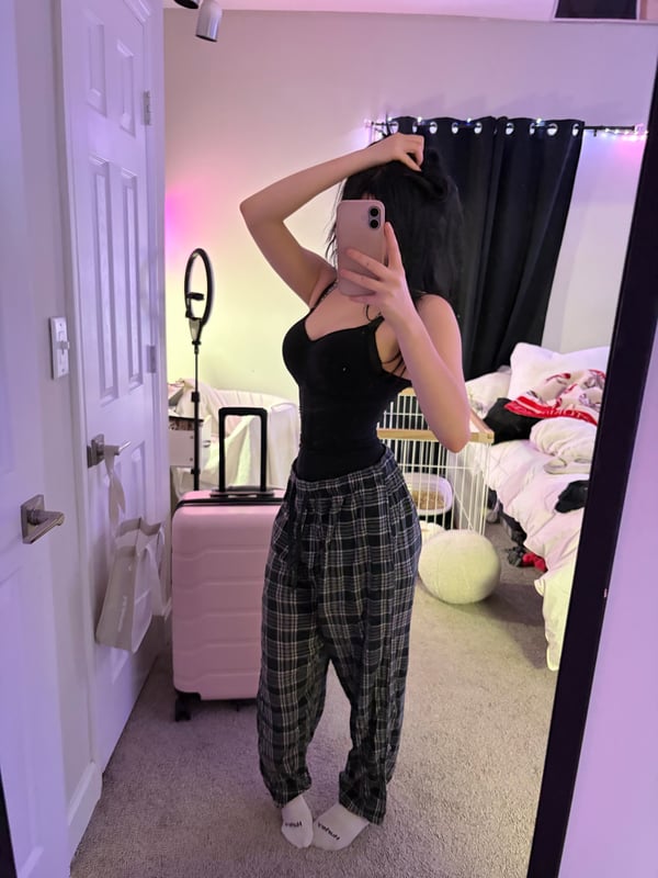 A person standing in a room wearing a black crop top and low-rise jeans, showing a navel piercing, with a wall outlet in the background.
