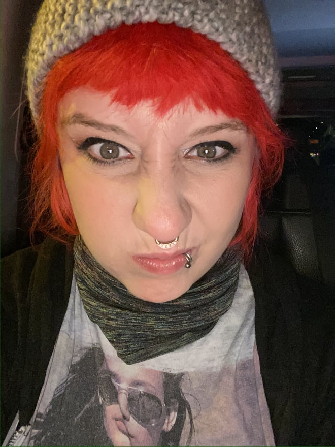 A woman with bright red hair and a knitted hat pouts at the camera, wearing a scarf and graphic t-shirt.