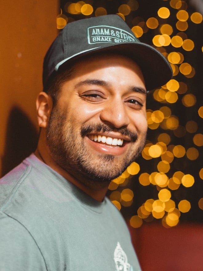 A smiling man wearing a cap and casual shirt, posing with warm lighting and bokeh lights in the background.