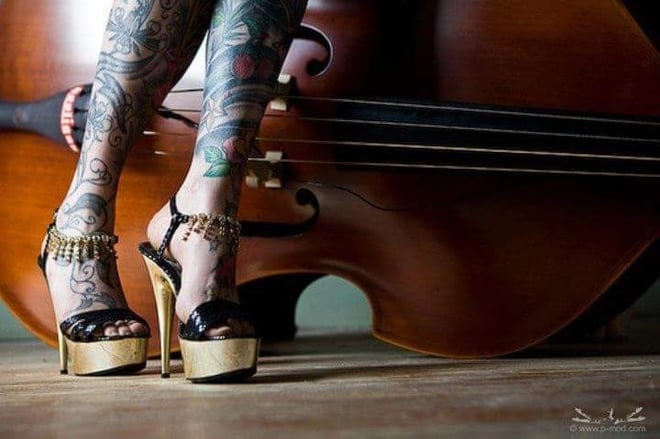 Close-up of a person's tattooed legs in high-heeled sandals positioned next to a large wooden double bass.