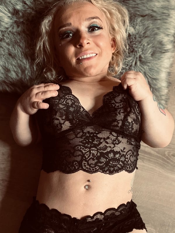 A petite blonde woman with tattoos and piercings smiling while lying on a fur blanket, dressed in black lace lingerie.