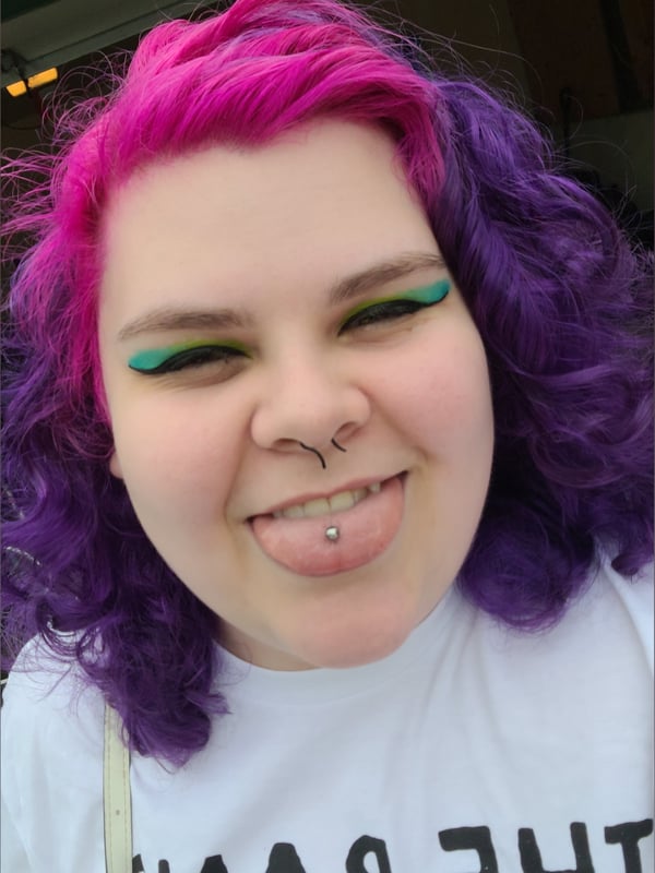 Young woman with vibrant pink and purple curly hair, green makeup, and piercings, smiling playfully while wearing a white shirt.