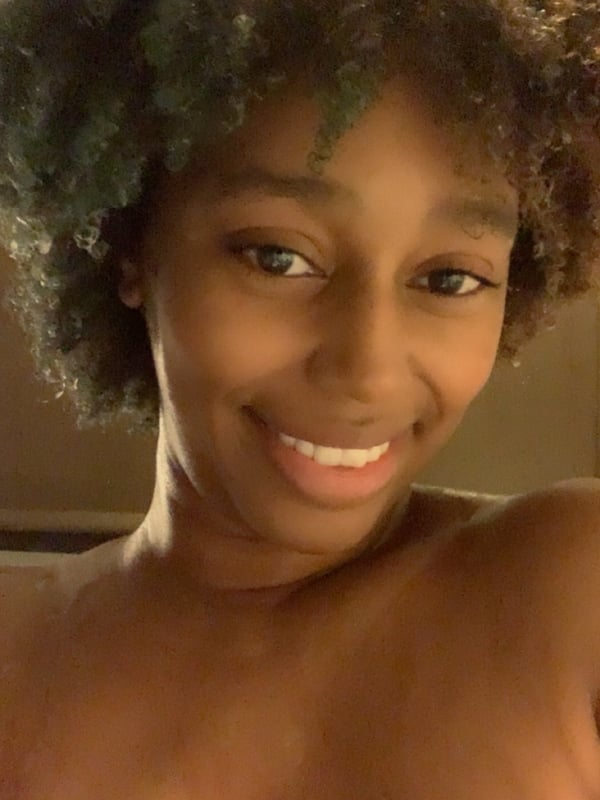 A smiling woman with curly hair taking a close-up selfie, showcasing her face and upper body in a casual indoor environment.
