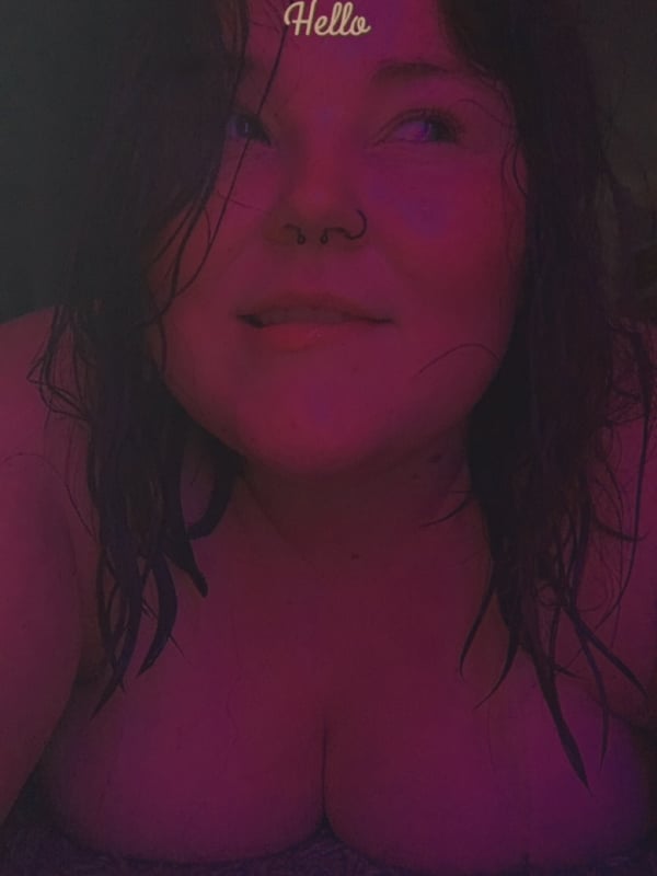 Close-up portrait of a young woman with wet brunette hair, smiling at the camera with 'Hello' text on her forehead, in a dimly lit indoor setting.