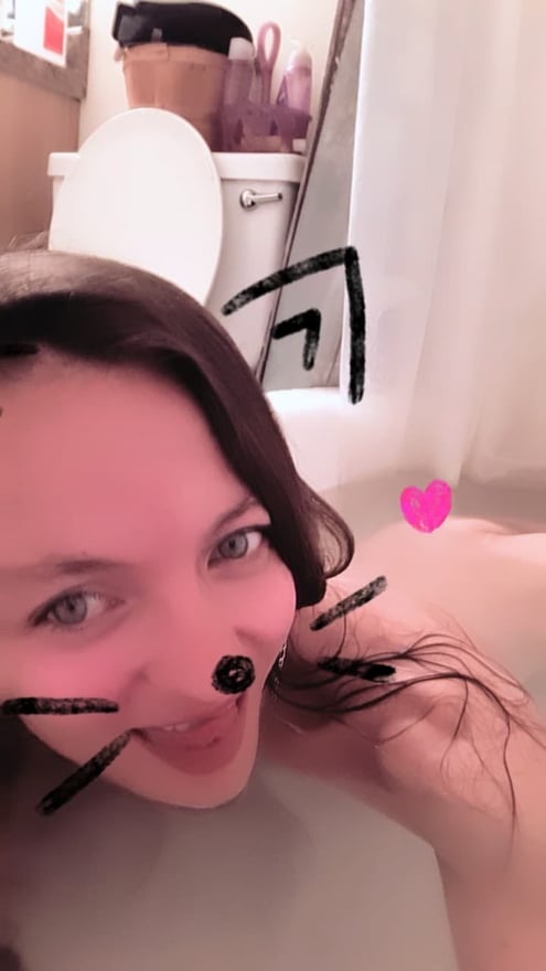 Woman with long dark hair making a playful cat face with drawn whiskers, smiling in a bathroom setting near a bathtub.