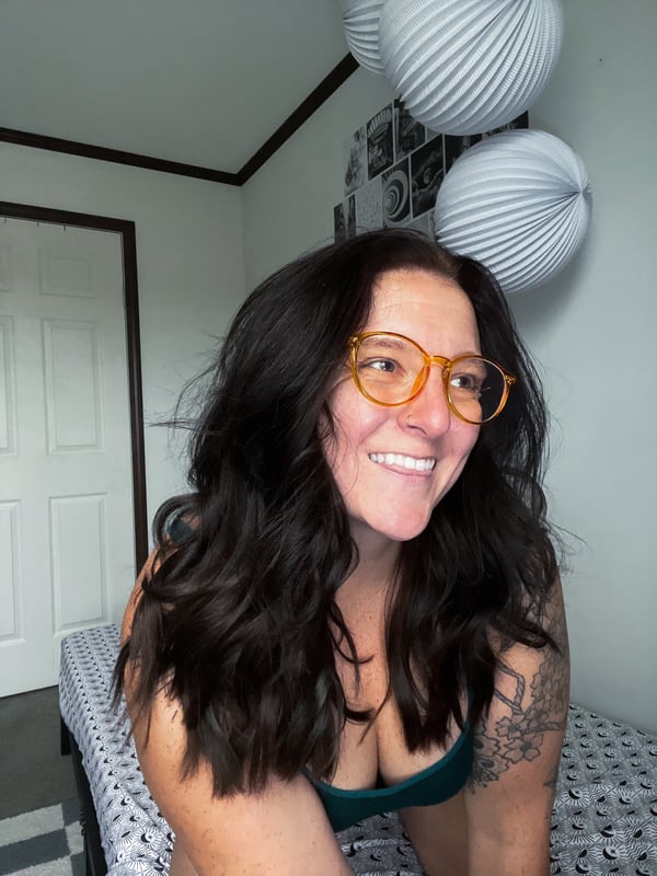 A smiling woman with long dark hair and orange glasses posing indoors near hanging paper lanterns, wearing a green top.