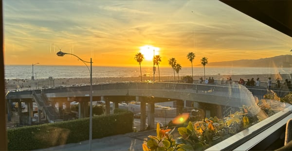 Scenic sunset view over a coastal bridge with palm trees, ocean, and city lights from an indoor window overlooking the Pacific Coast Highway.