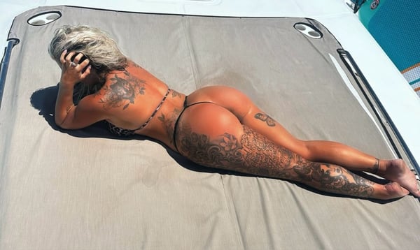 A woman lying on a boat deck in a bikini, posing relaxed with tattoos visible against a sunny outdoor background.