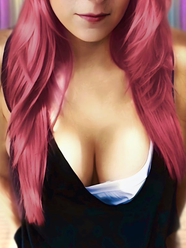 Woman with long pink hair wearing a low-cut black top, posing confidently in front of a purple-toned garden background.