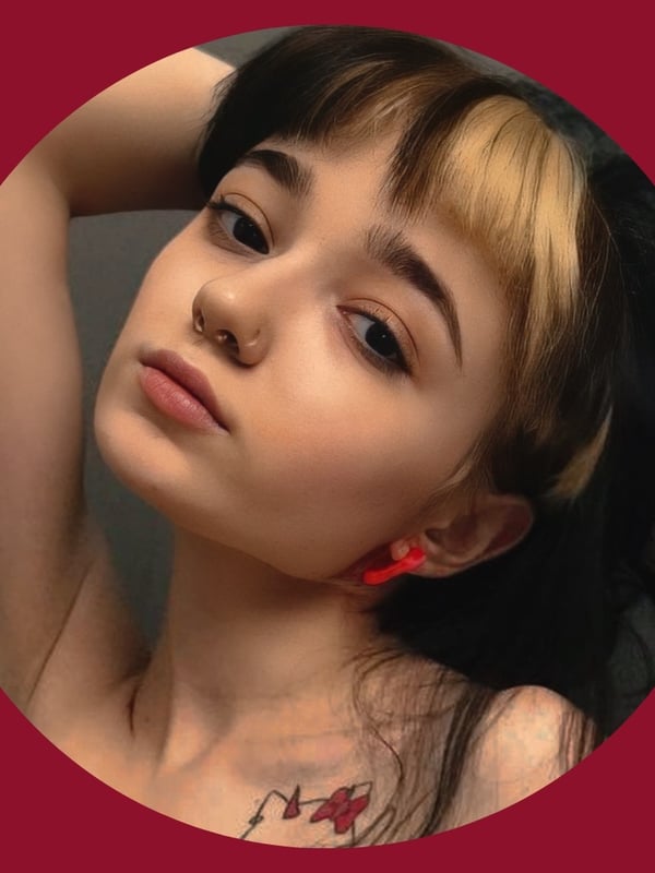 Close-up portrait of a young woman with dyed black and blonde hair, piercings, and a necklace against a red background.