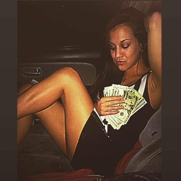 A young woman sitting in a car, holding cash, dressed in a tank top and shorts, with a relaxed pose.