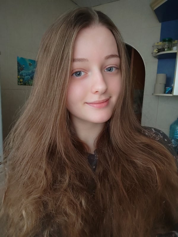 Young woman with long wavy brown hair smiling at the camera in a casual indoor setting with household items in the background.