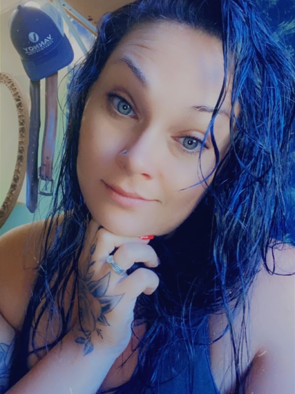 Close-up portrait of a woman with wet dyed blue hair, blue eyes, and tattoos, smiling softly indoors with decorative shelves in the background.
