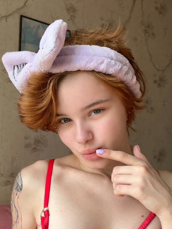 Close-up of a young woman with red hair and a pink bunny ear headband, pouting playfully with a finger to her lips in a bedroom setting.