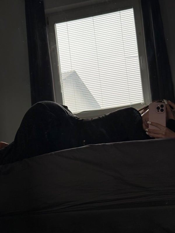 A person lying on a bed in dark clothing, holding a phone, with a window and curtains in the background.