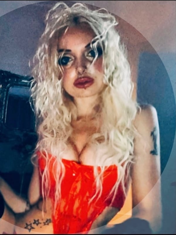 Close-up of a blonde woman with heavy makeup, wearing a red corset top, framed in a circular image.
