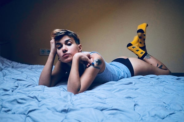 A person with short hair reclining on a bed in a tank top and shorts, wearing yellow socks, in a warmly lit room.