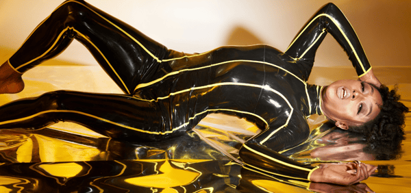 A woman posing dynamically on a reflective golden surface while wearing a shiny black outfit with yellow accents.