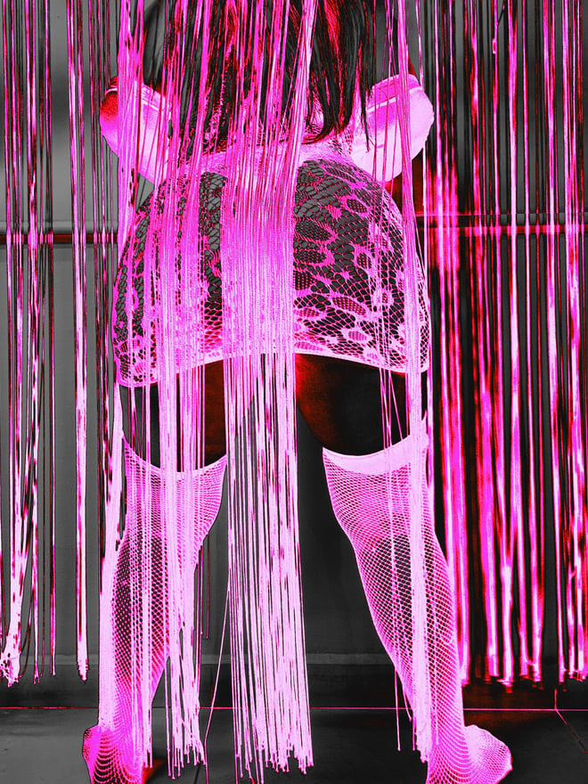 Woman standing with back view in lace lingerie and stockings against pink beaded curtains in a vibrant indoor setting.