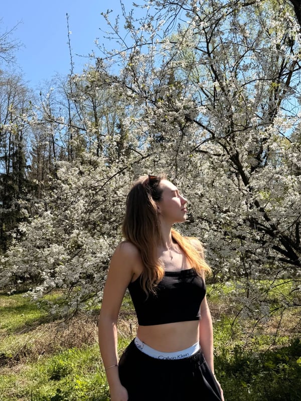 Young woman with long hair posing sideways in a black crop top and sweatpants amid blooming cherry trees under a blue sky.