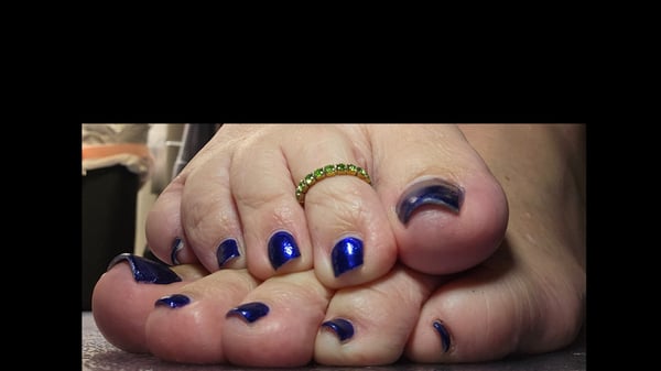 Close-up view of bare feet with dark blue nail polish and a green ring on one toe, positioned near a dark surface.
