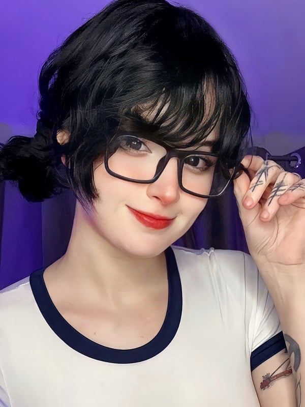 Young woman with glasses and dark hair styled in pigtails, smiling playfully against a purple background.