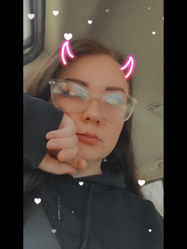 A young woman with glasses and heart-shaped horns filter, wearing a hoodie, posing playfully indoors with heart effects.