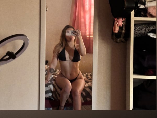 A woman with long blonde hair taking a mirror selfie in a black bikini, sitting on a bed in a bedroom with a ring light and clothing nearby.