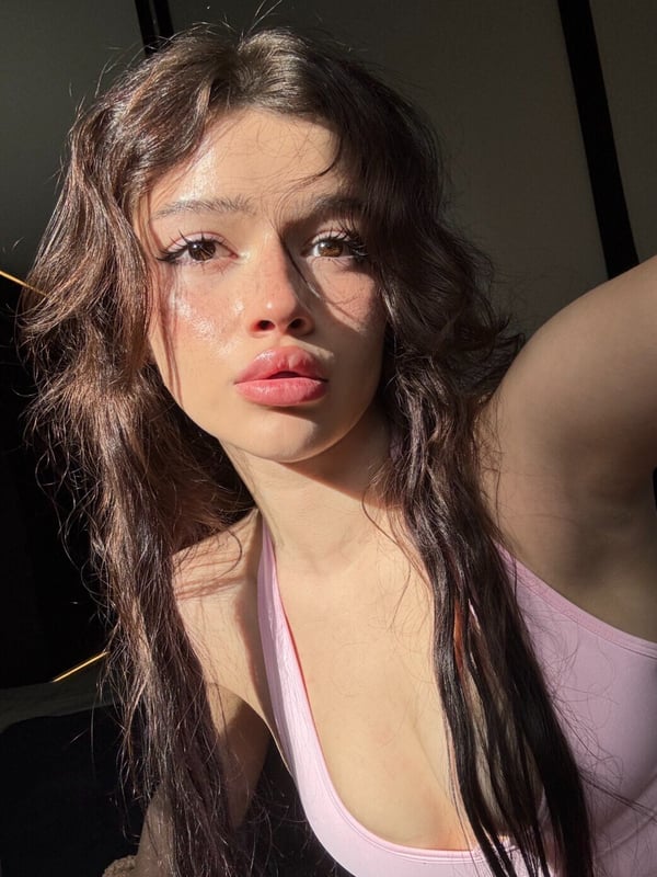 Close-up selfie of a young woman with long dark hair wearing a pink top, taken in natural lighting.