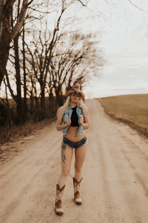 Woman wearing a cowboy hat, open denim jacket, crop top, shorts, and boots posing confidently on a rural dirt road lined with bare trees and fields.